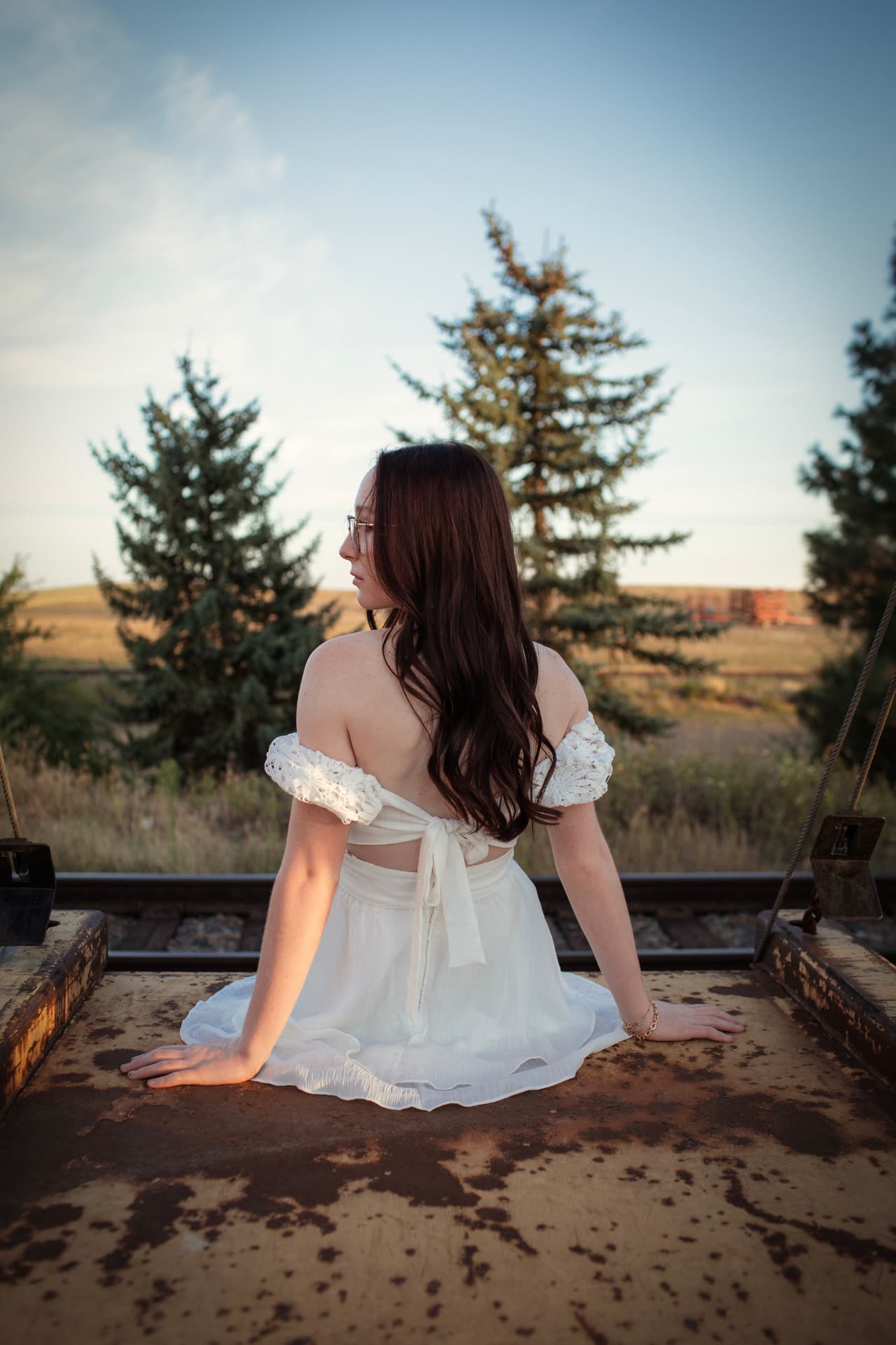Senior in a white dress seated on a rail car, looking toward the pines
