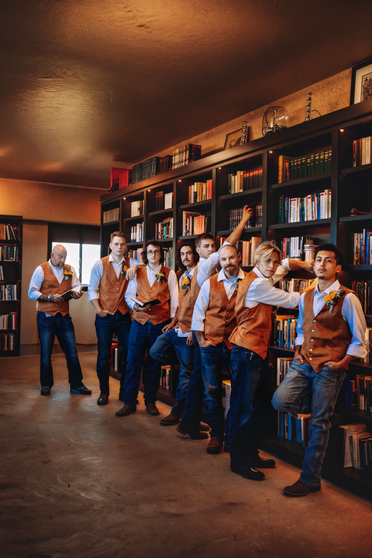 Groomsmen in rust-colored vests leaning against a library bookshelf before the ceremony