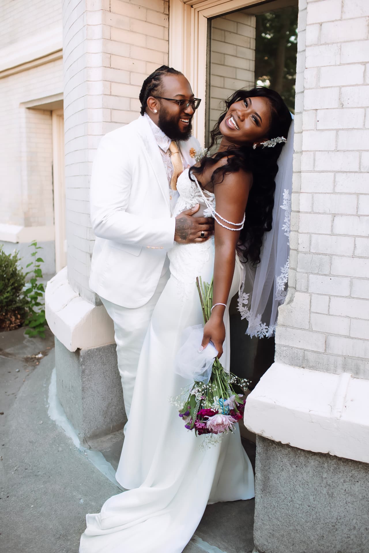 Bride and groom laughing together by a white brick building, bouquet in hand