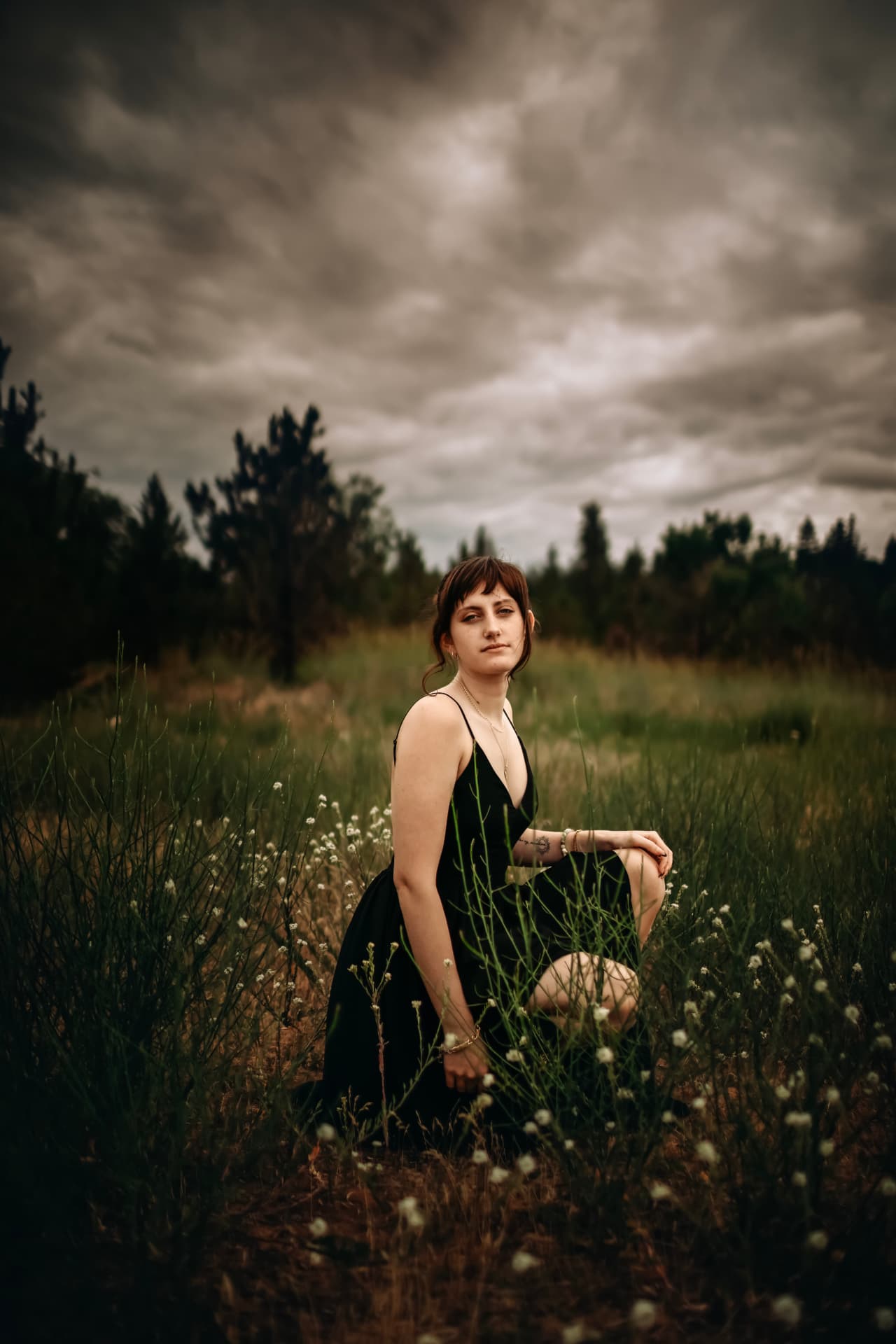 Senior in a black dress crouching in a wildflower meadow under dramatic clouds