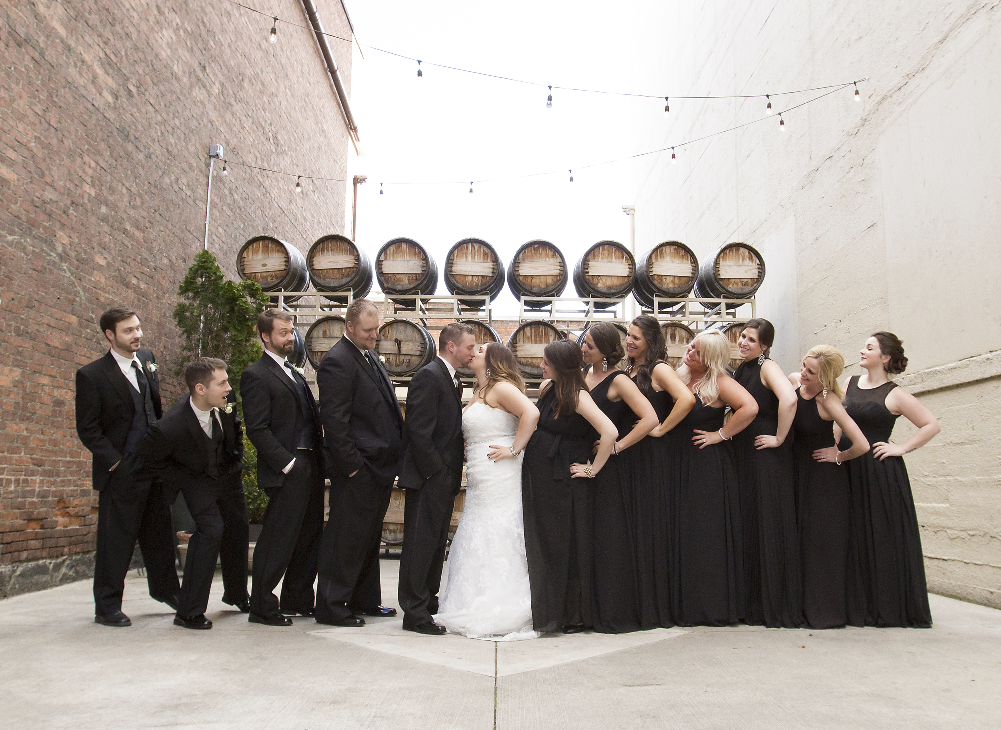 Bridal party in black gowns and suits beside wine barrels in a brick alley