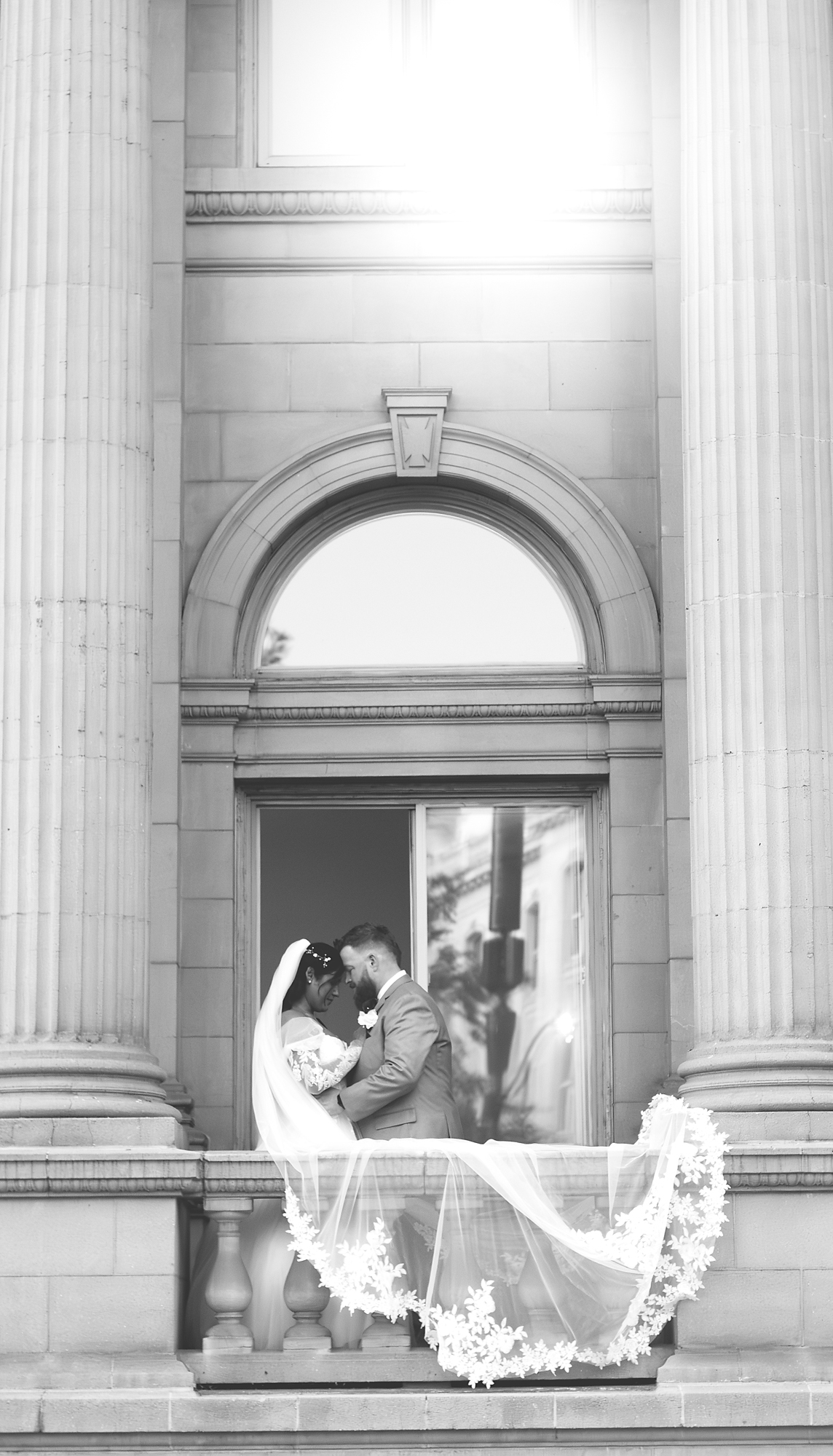 Black and white photo of a bride and groom on a courthouse balcony, veil trailing over the railing
